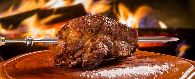 Imagem de peça de carne suculenta em um espeto de churrasco, recém-retirada do fogo. Ao fundo, chamas de fogo.