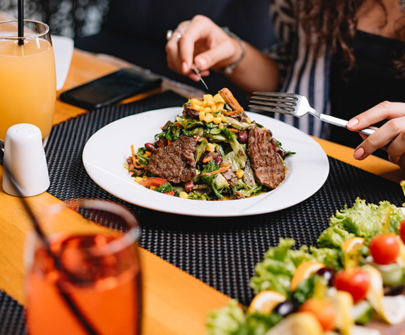 Zoom em um prato com dois bifes de carne, salada e grãos de milho cozido. Ele está apoiado em uma mesa de madeira.